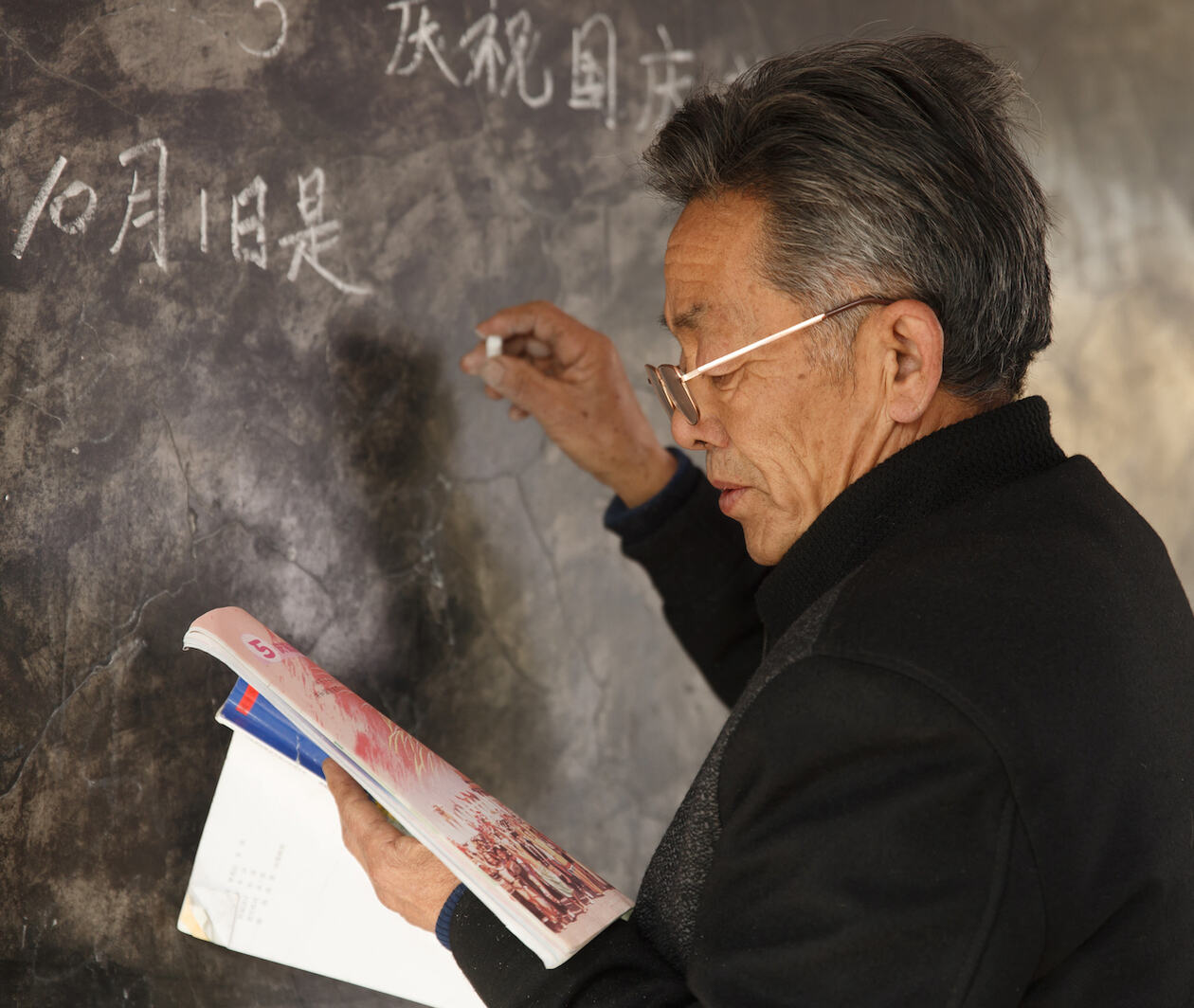 Elderly male teacher reading from textbook while writing Chinese characters on black board.