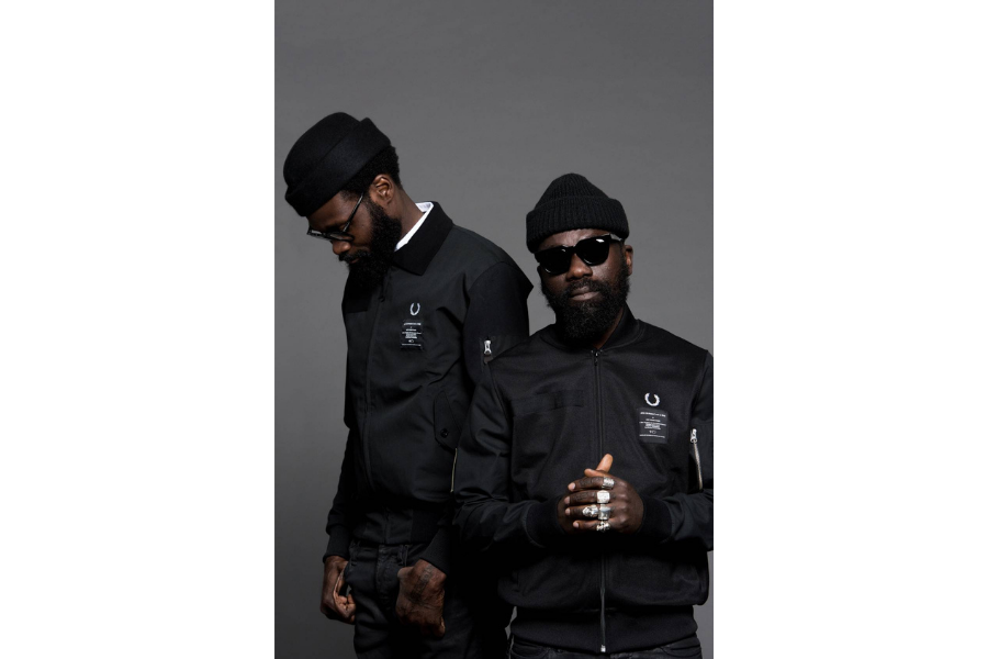 Sam and Shaka wearing ACFxFred Perry bomber jackets, hats and glasses. Sam (right) is facing the camera. Shaka (left) is looking down.