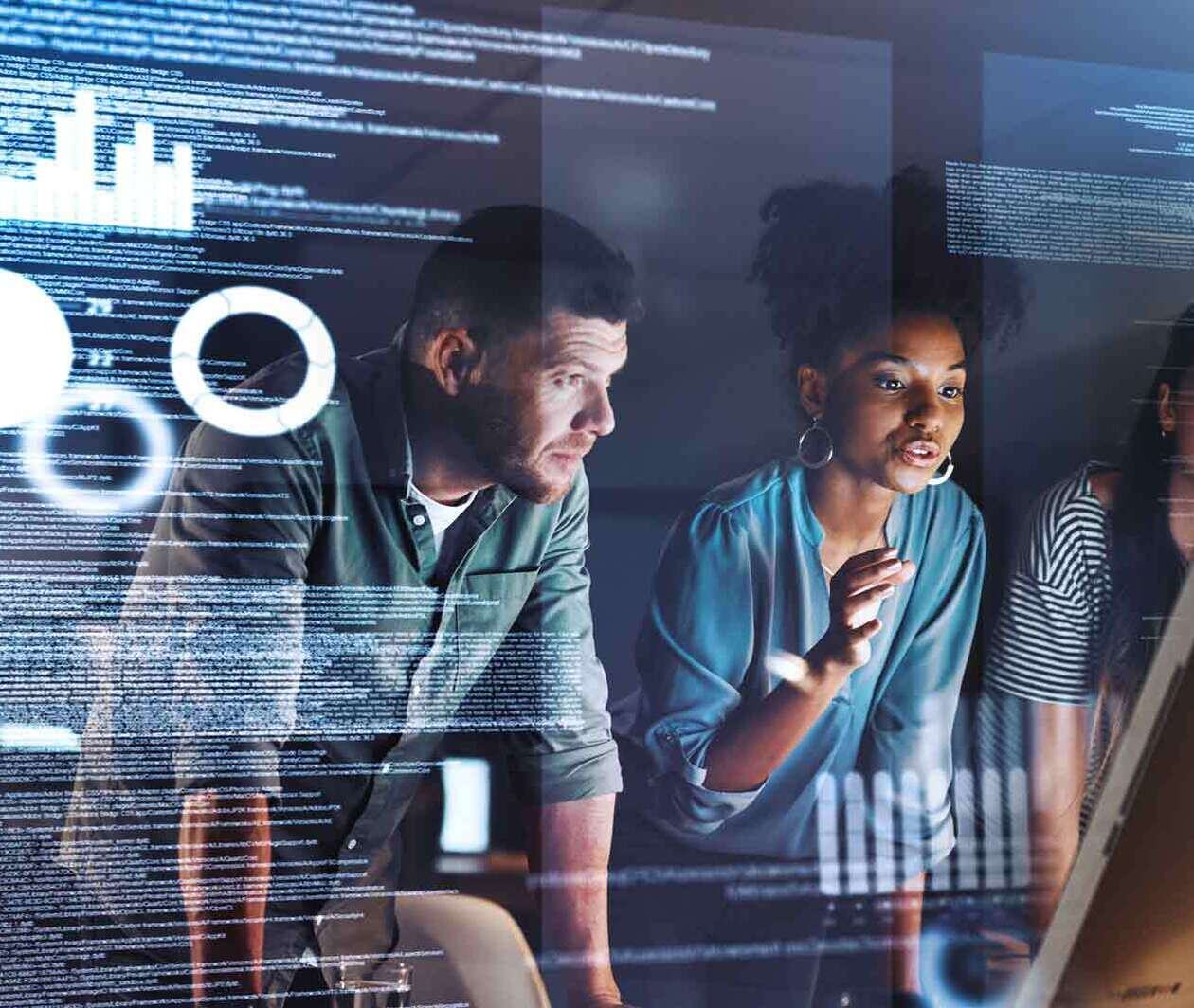 a man and two women stand side-by-side looking intently at a computer monitor whilst surrounded by reflected imagery of computer coding and graphs