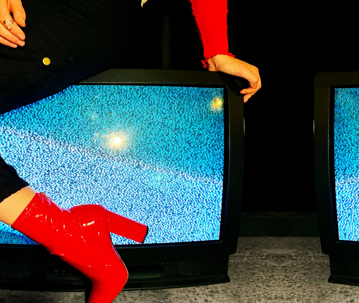 A woman in red high-heeled boots sitting atop some old TV screens showing static