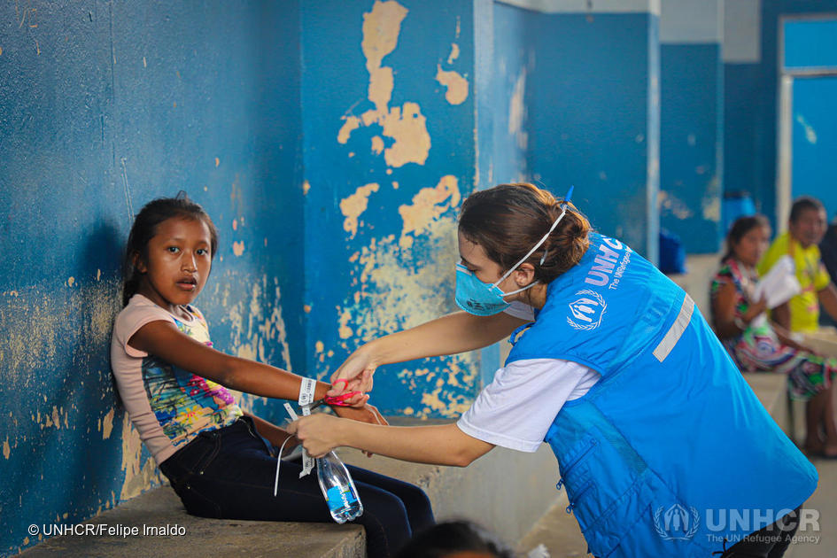 Young girl sitting on a bench inside a building with a woman wearing a mask from UNHCR putting a label around the girls wrist.