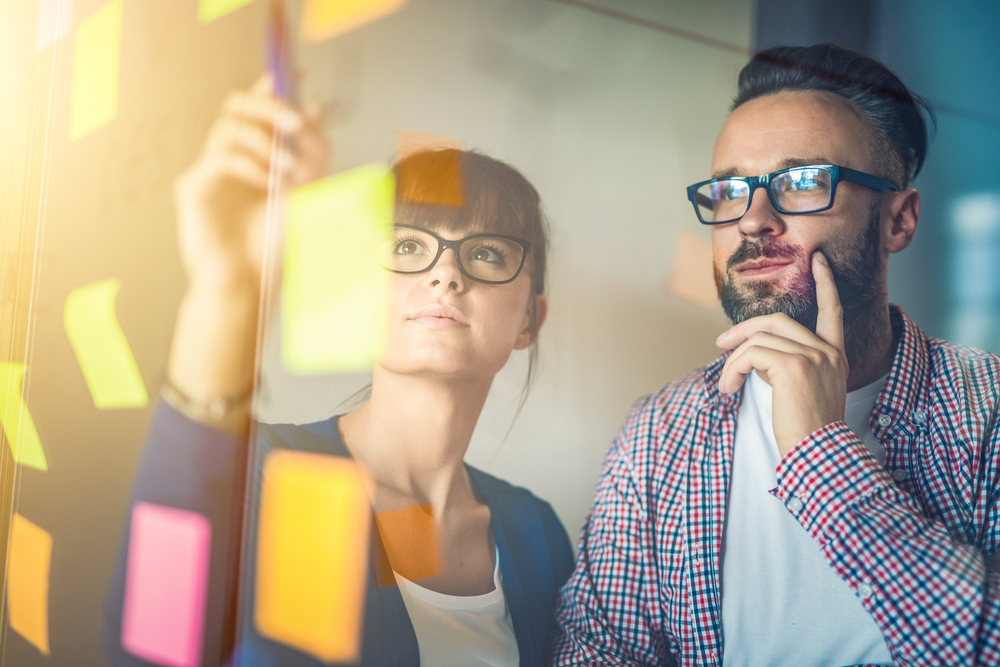 Two people contemplate a range of post-it-note memos stuck on to a glass wall.