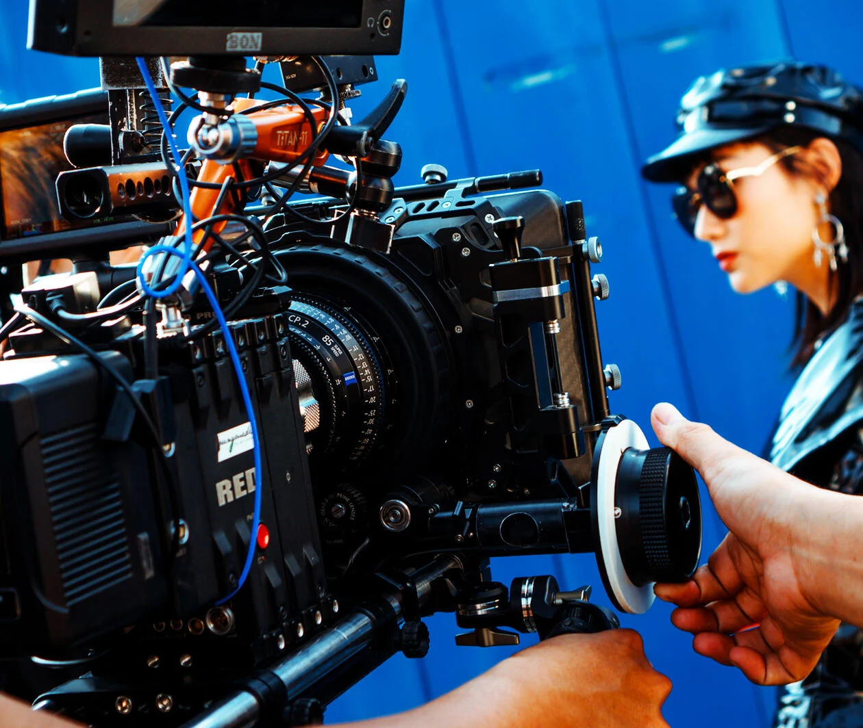 Close up of a camera and operator filming a young lady dressed in a black leather jacket and hat; wearing sunglasses.