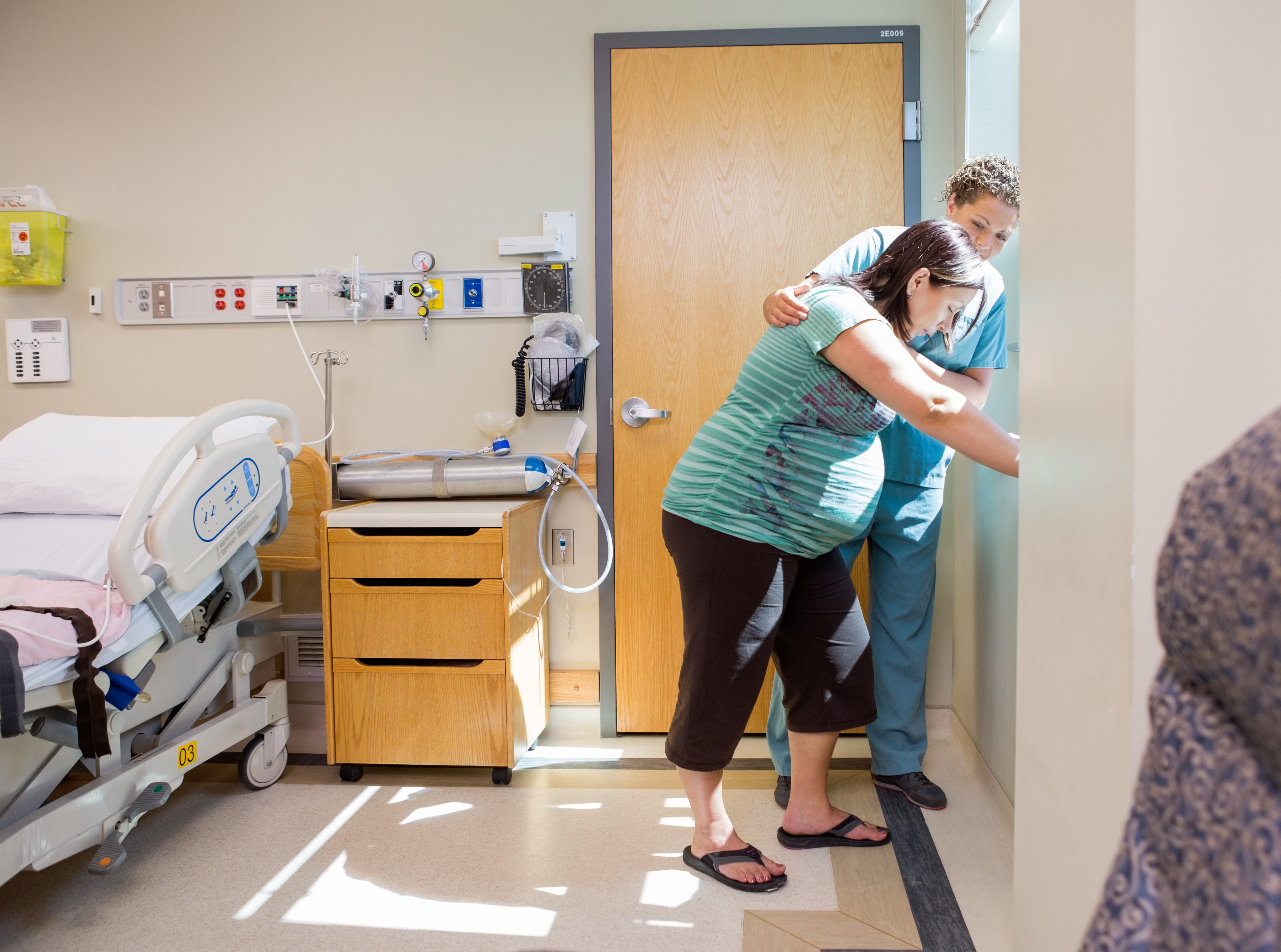 A pregnant woman leaning against the window of her hospital room being supported by a midwife