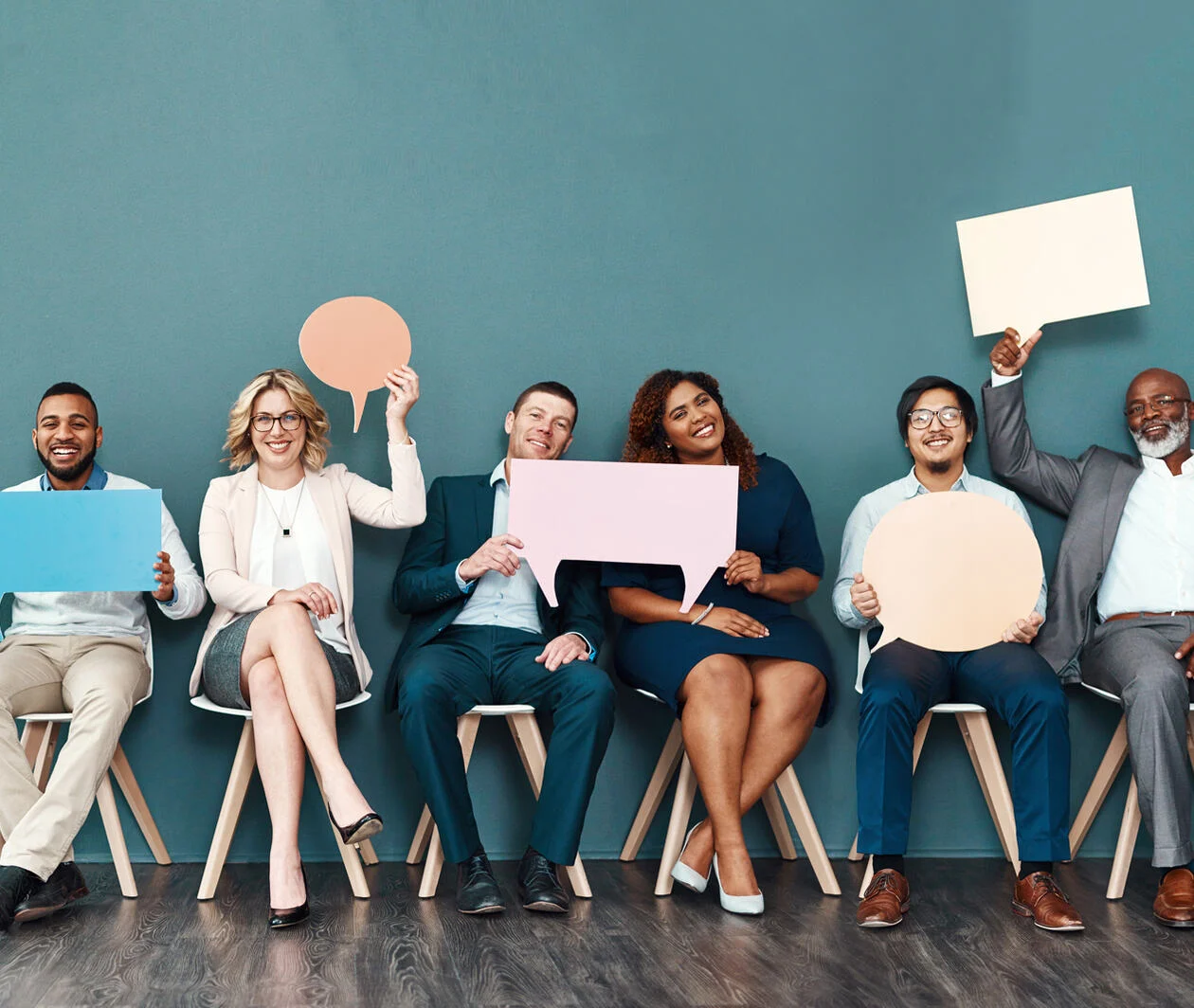 Shot portrait of a diverse group of business people with men and women holding up speech bubbles while they wait in line in front of a blue wall.