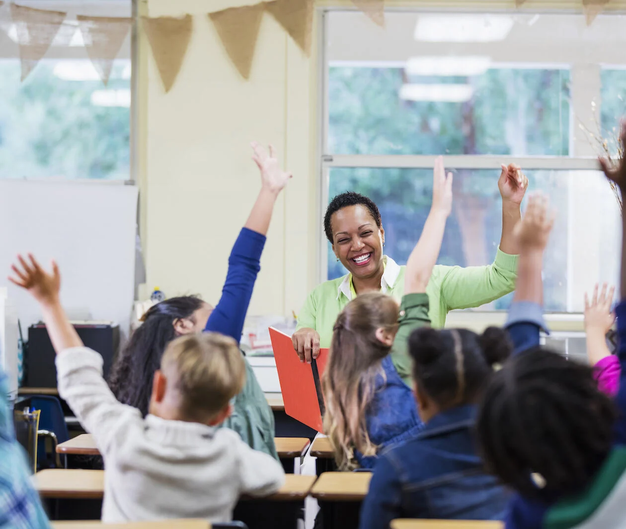 Teacher smiling at a group of primary age students with their hands up