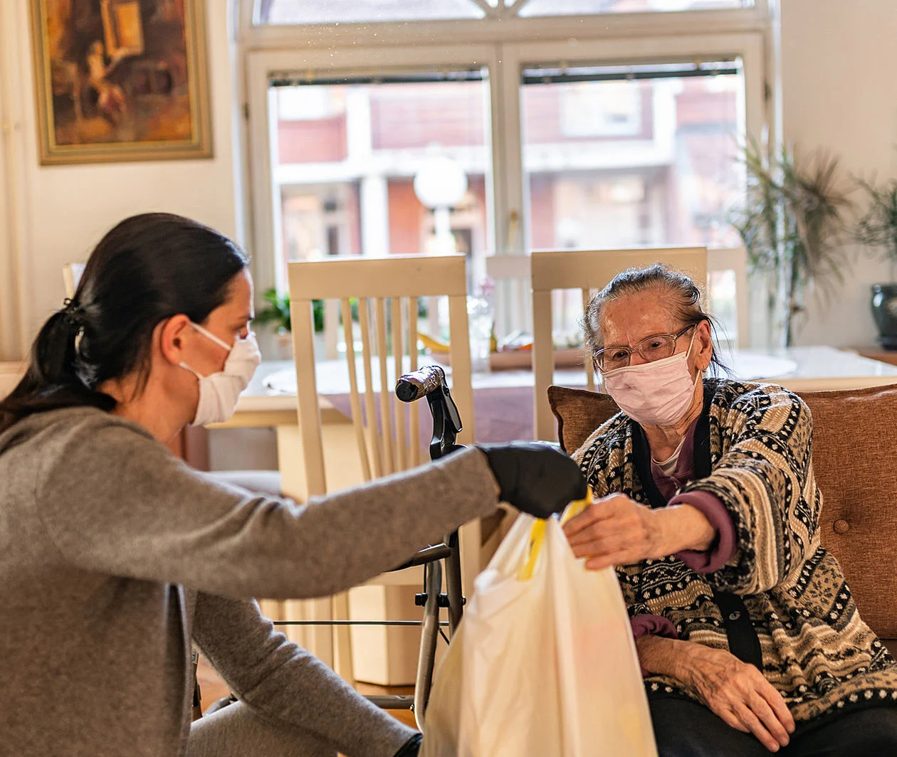 Two women wearing face masks sat down. The younger woman is handing the older woman a plastic bag