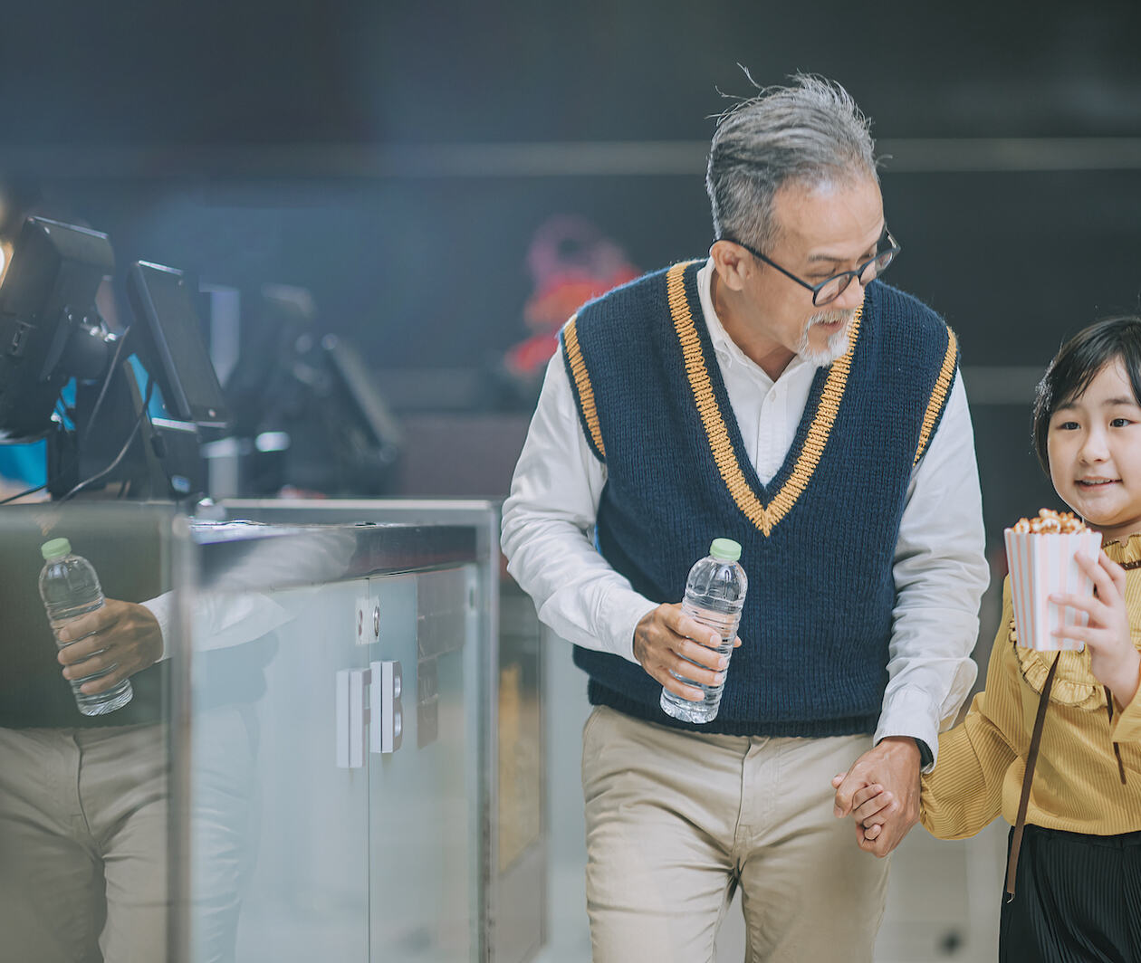 A Chinese grandfather holding his granddaughters hand, he is also holding a bottle of water and she holding a cup of popcorn. They are in conversation as they are entering the movie theatre, having just brought movie tickets