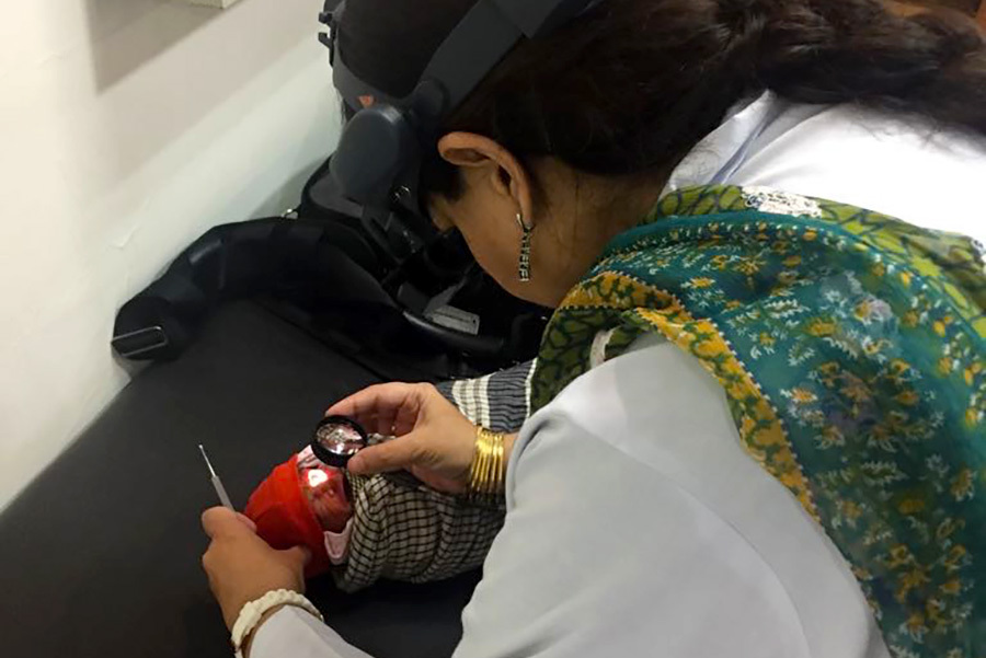 Ophtalmologist, wearing an ophthalmoscope on her head, is bent over the baby and holding a lens close to the baby's eye