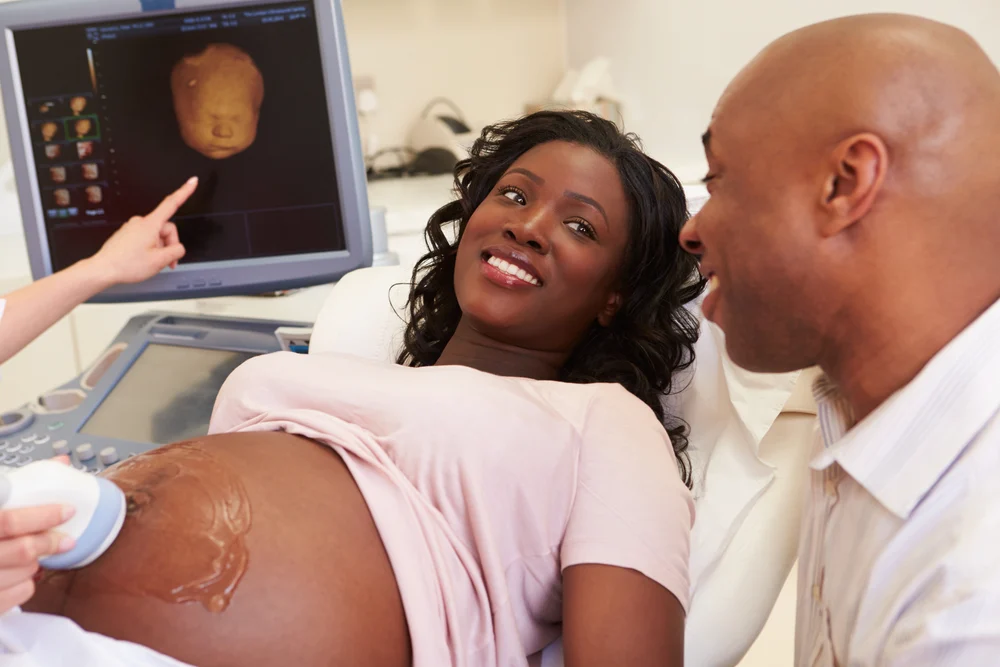 A pregnant woman lies on an examination table, while an ultrasound scan is being performed on her abdomen. Her unborn child is shown in the computer monitor in the background and she is supported by a partner.