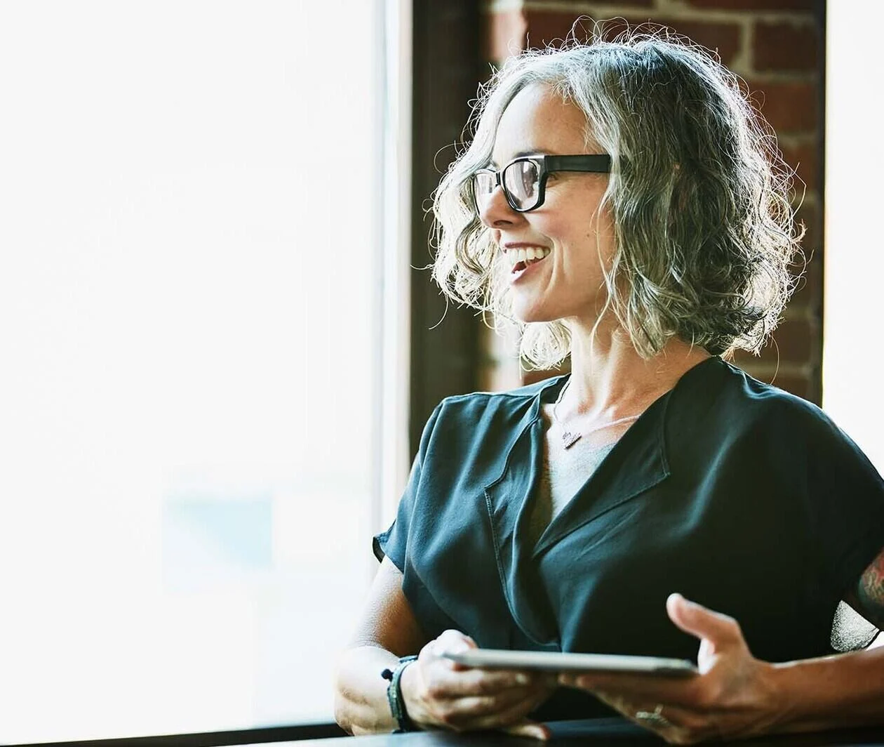 A woman has short grey hair and glasses and is sat in an office holding a tablet device.