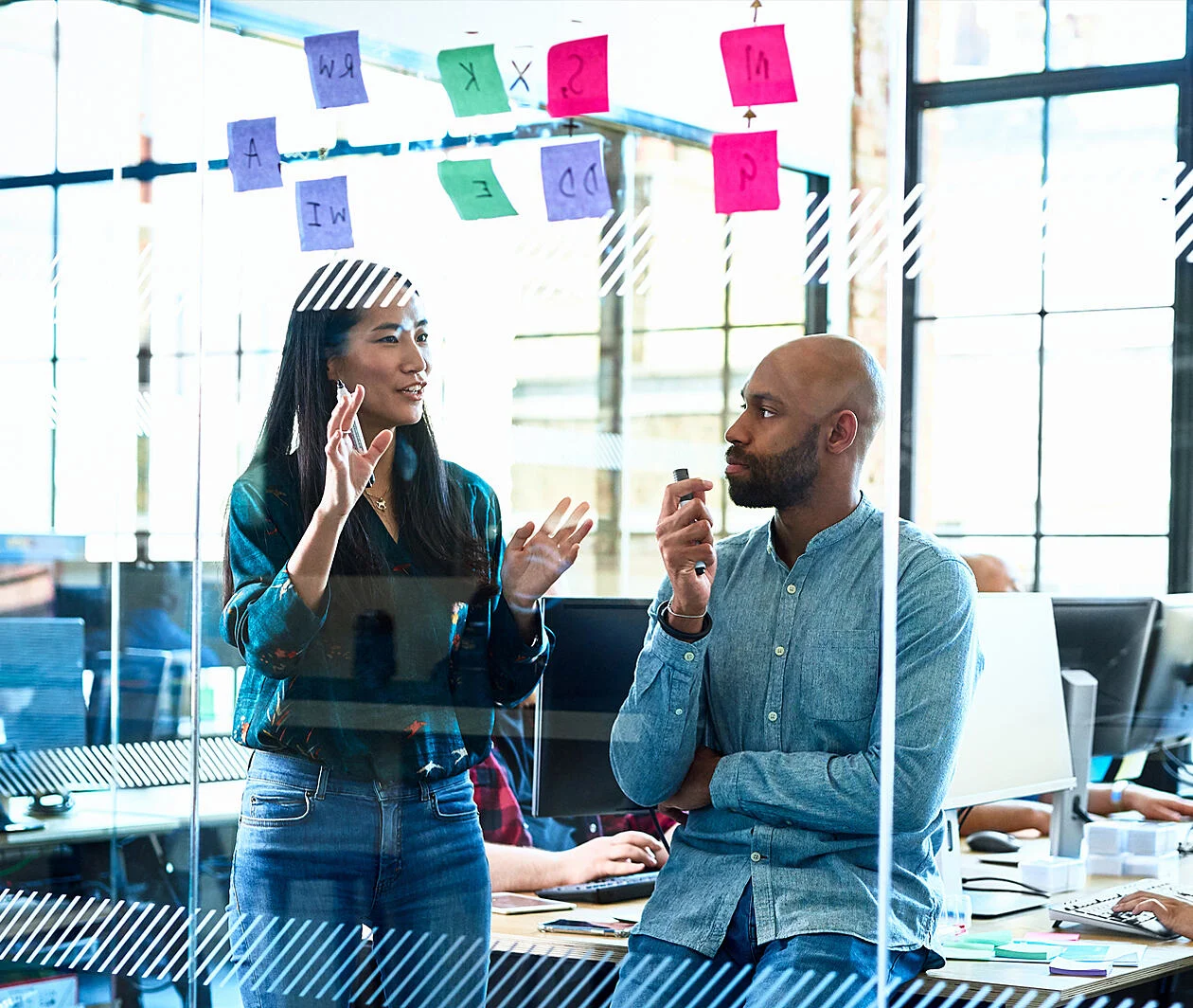 Two people talking in front of a glass wall with post it notes on it.