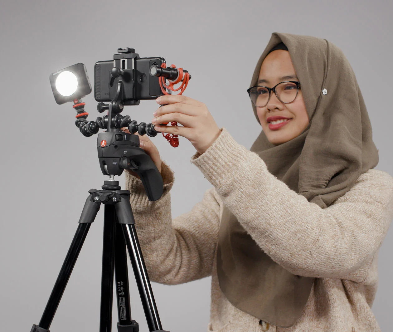 An image of a woman wearing a head scarf and jumper. She is adjusting a smartphone set up for filming atop a tripod.