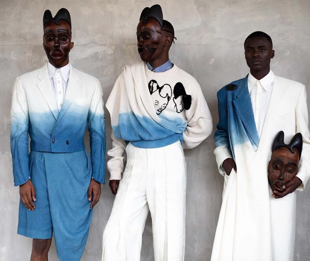 Three men posing in front of a concrete wall wearing blue and white garments and wooden masks.  