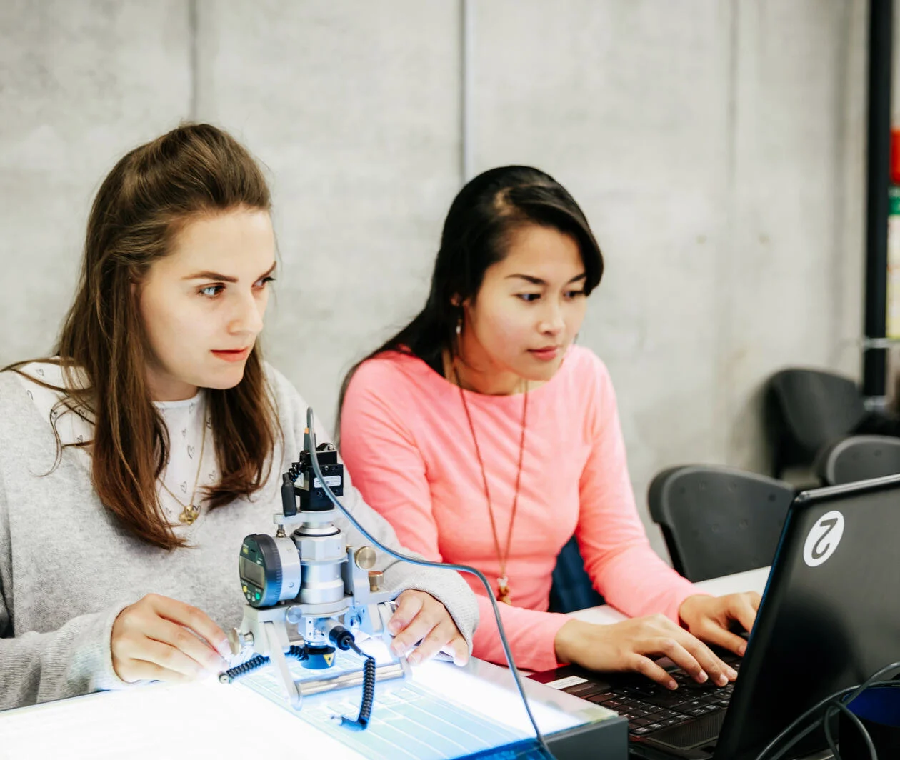 two young women using a computer to operate a robot. One in a pink jumper, the other in a yellow shirt