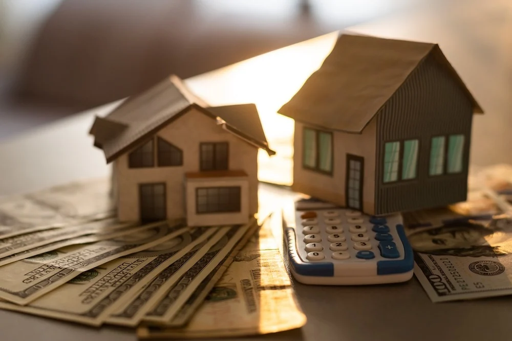 Close up of two model houses sitting on calculator and fans of cash