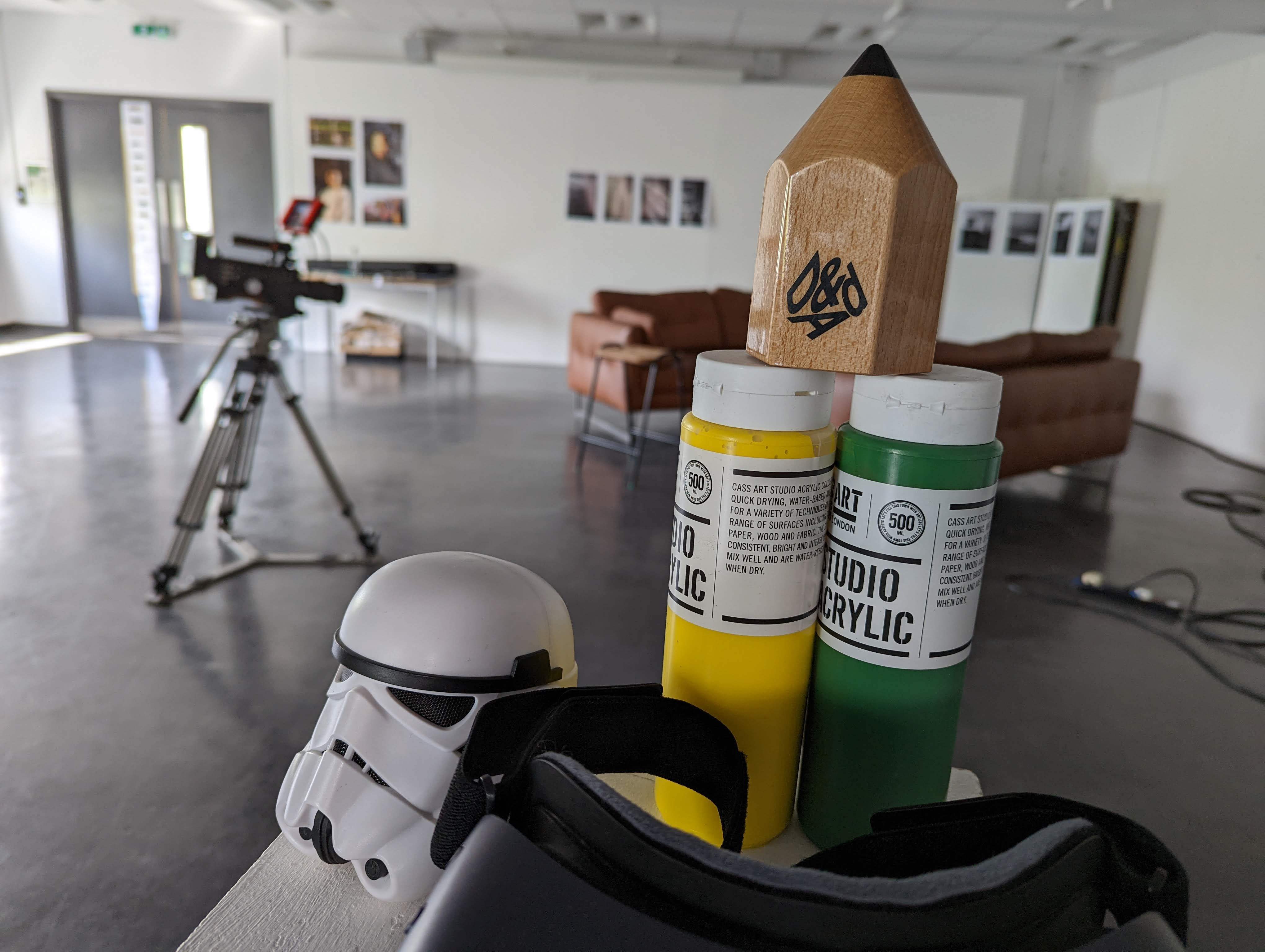 On a white plinth there is a black VR headset, a Storm Trooper helmet, two bottles of paint in green and yellow, and an Advertising and Design Award. In the background there is 2 brown sofas and a camera set up.