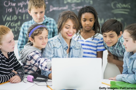 A female teacher with 3 pupils on either side. They are all looking towards a computer, the screen of which is facing away from the camera. One of the female pupils is pointing at the screen.