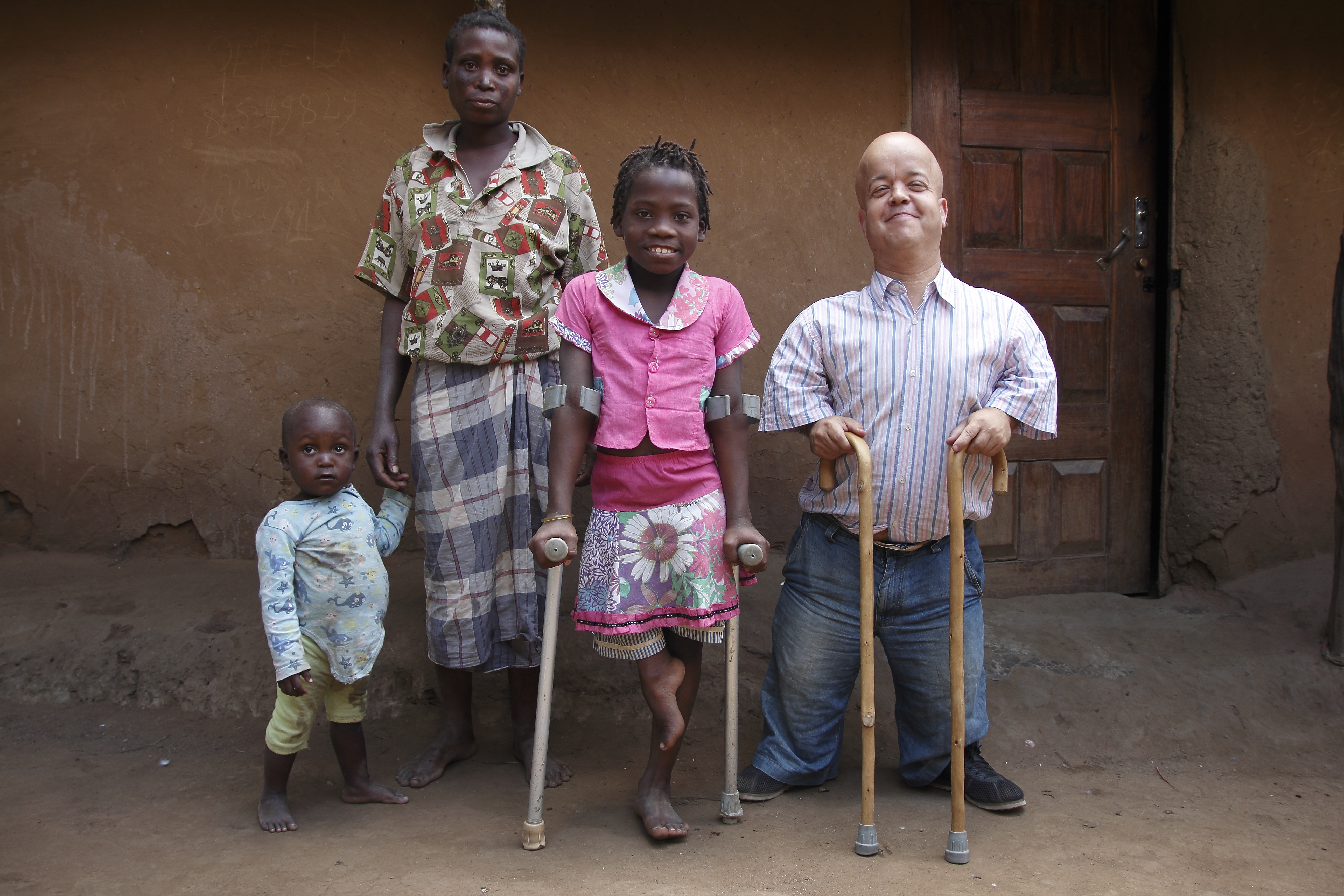 Image shows four people posing for camera, from left to right a child holding his mother’s hand, an older female child using crutches, and a man using walking sticks.