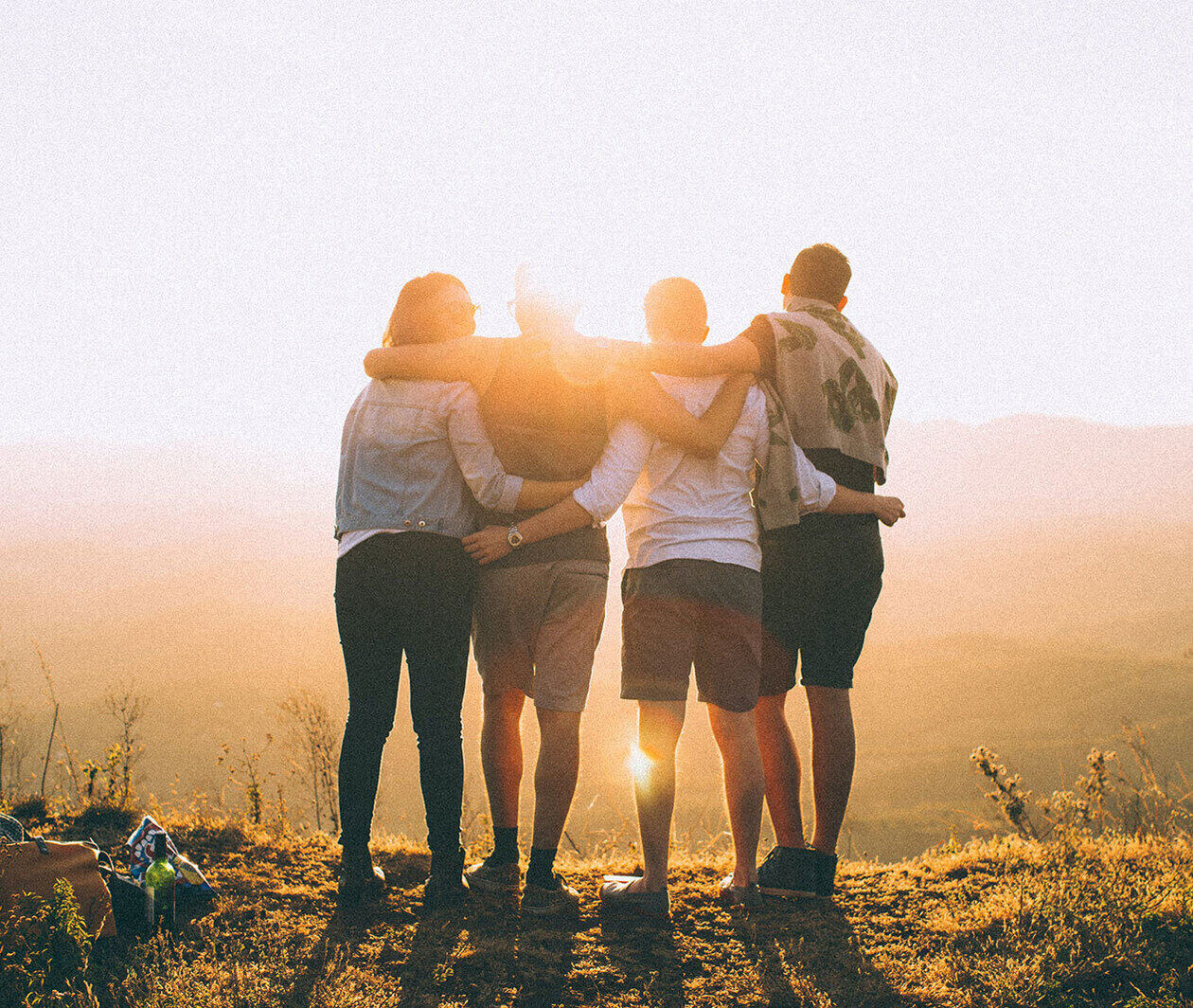 A group of young friends standing side-by-side hugging as they stare off into the sunset atop a hill