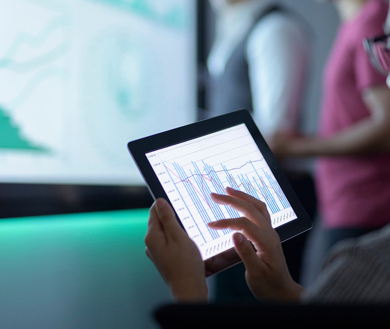 Someone working on a tablet with charts onscreen. A larger screen in the background with two people standing near also shows charts.