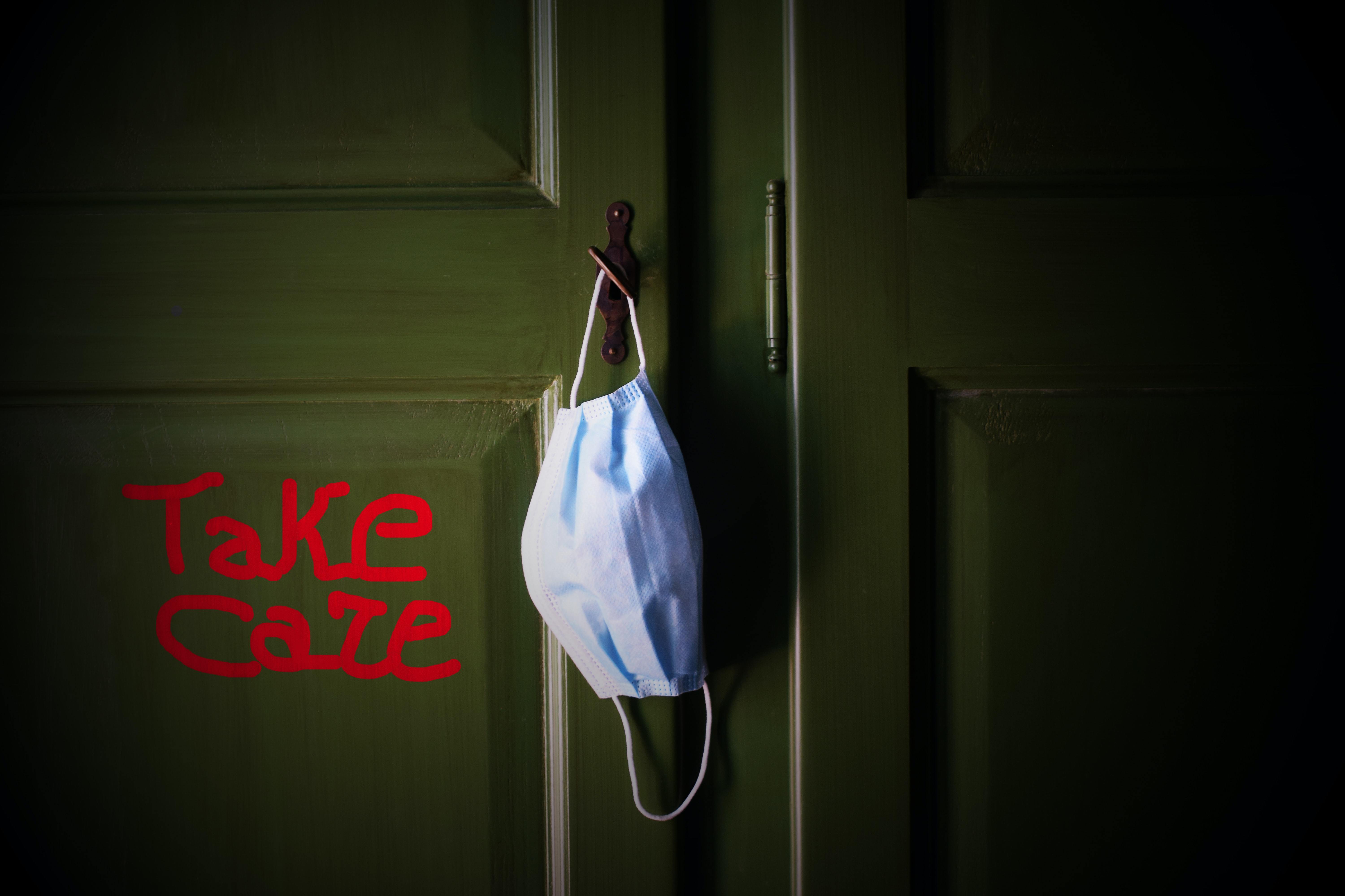 Take Care' written in red on a green door with a face mask hanging from the door handle.