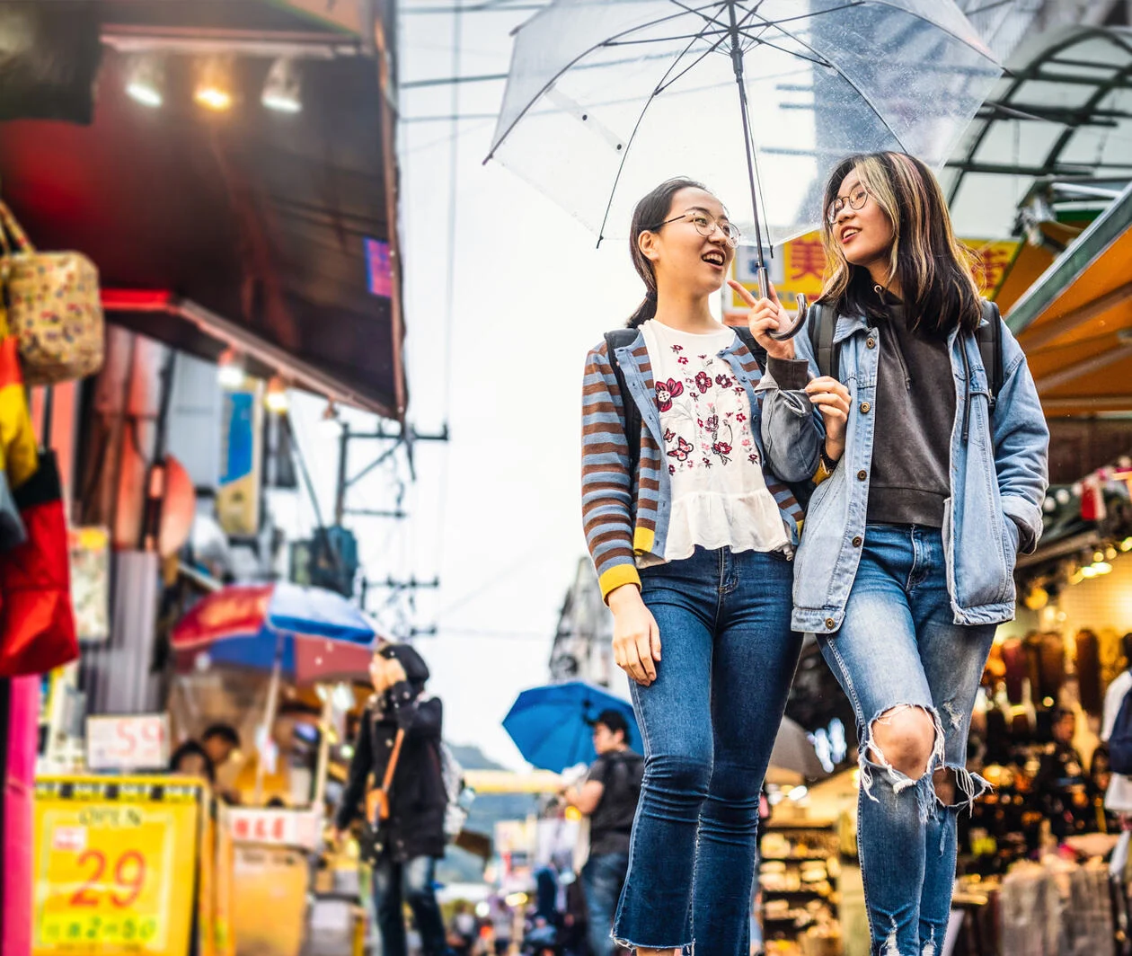 Two happy asian girls holding an umbrella and walking in a street market.