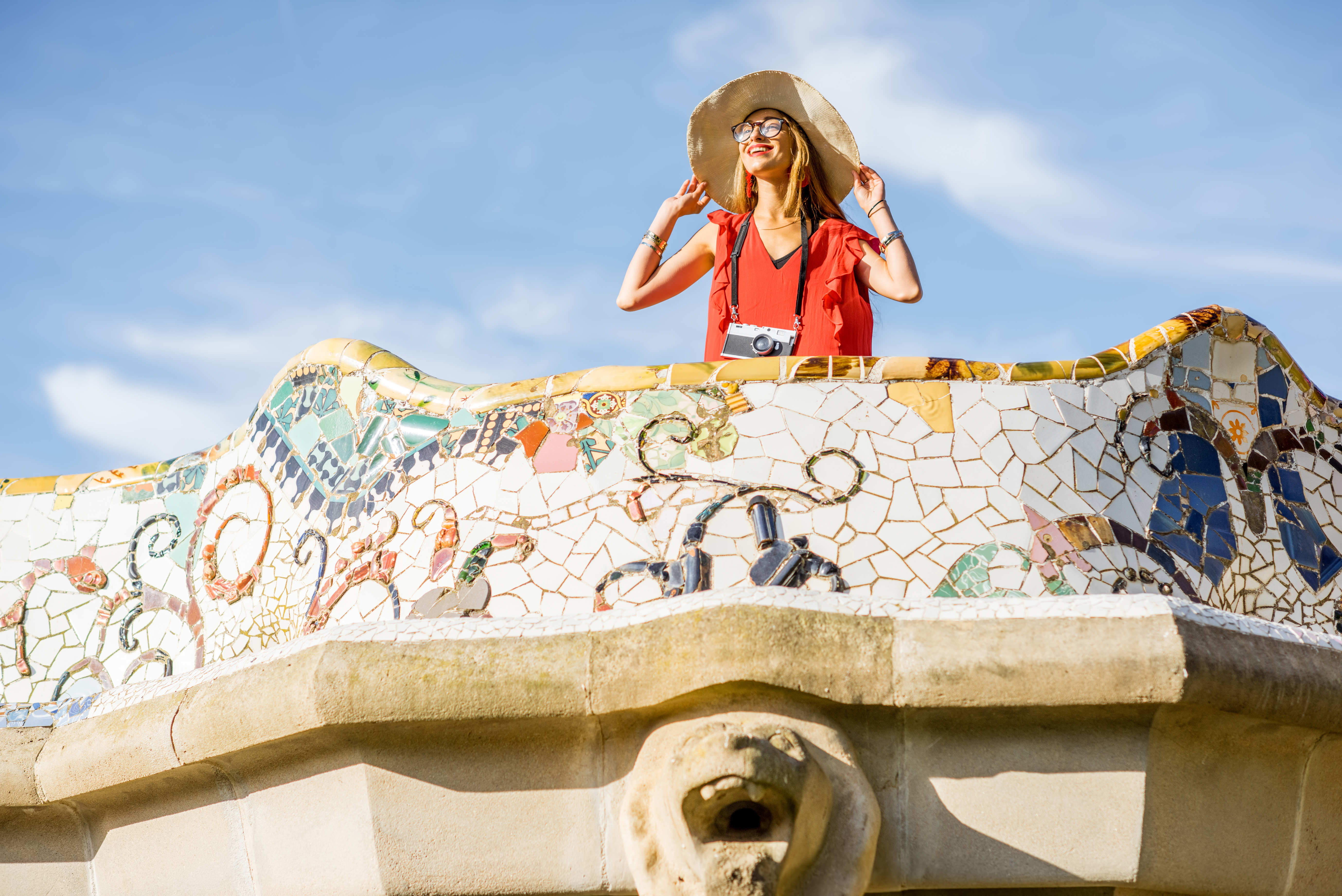 A woman standing on the beautiful terrace of Guell park in Barcelona. She is wearing a sun hat and is smiling. The sky behind her is clear with a few clouds.