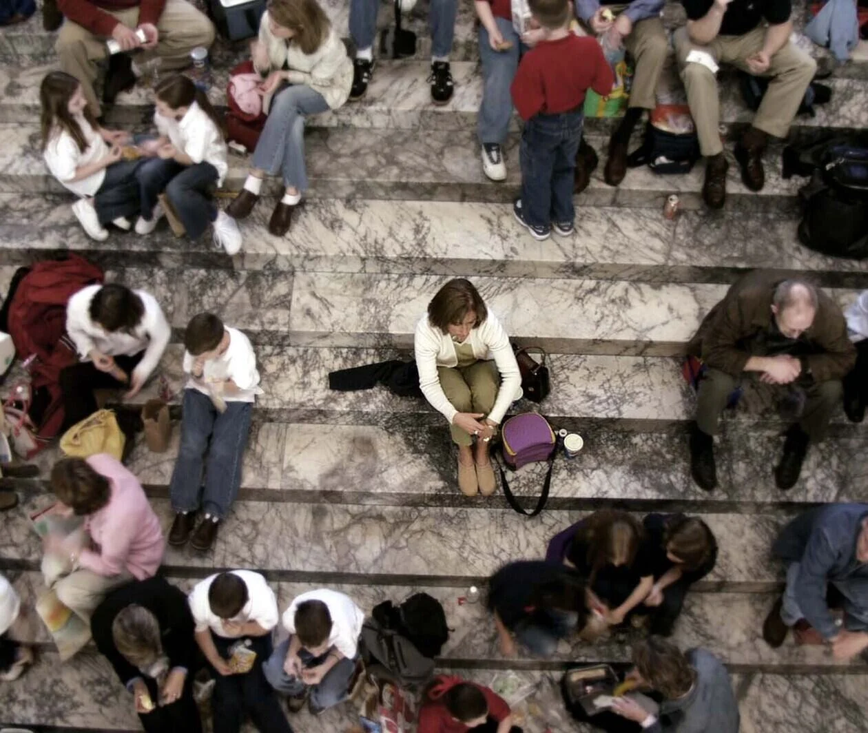 A woman sits on her own, isolated, on a flight of stairs surrounded by a large crowd of people.