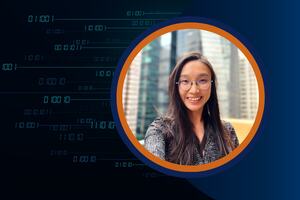 A professional woman with long dark hair and glasses smiles at the camera, with tall city buildings in the blurred background. She is framed in a circular border with an orange and navy blue ring.