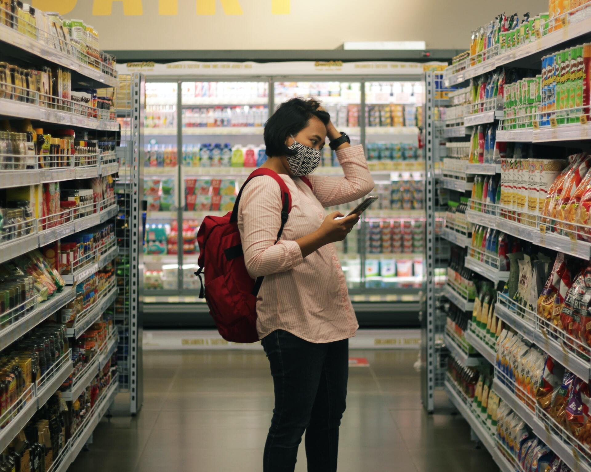 Photo of a woman standing in front of supermarket shelves with a phone, looking perplexed