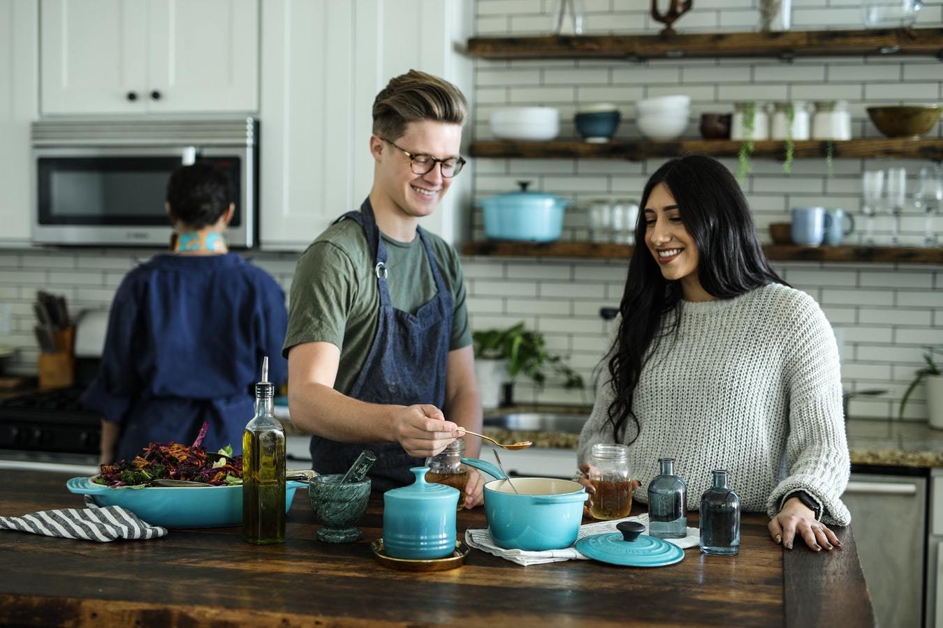 a smiling man wearnig an apron, standing near a smiling woman in a kitchen. Both are looking at a cooking pot.