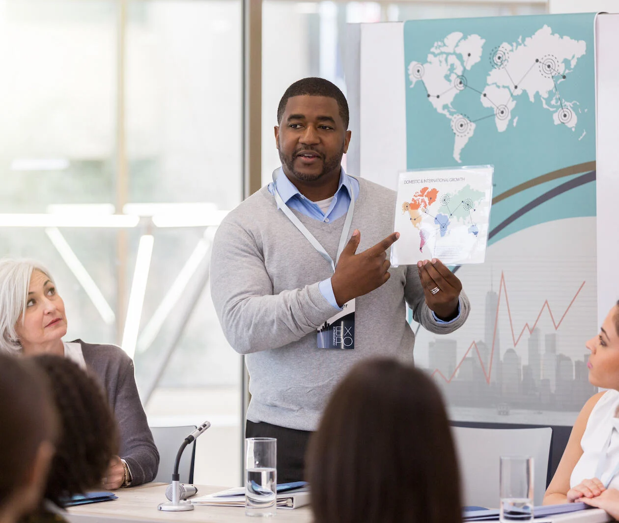 Male professional presenting a world map covered in statistics to colleagues around a desk. With a similar diagram behind him, and female colleagues either side of him.