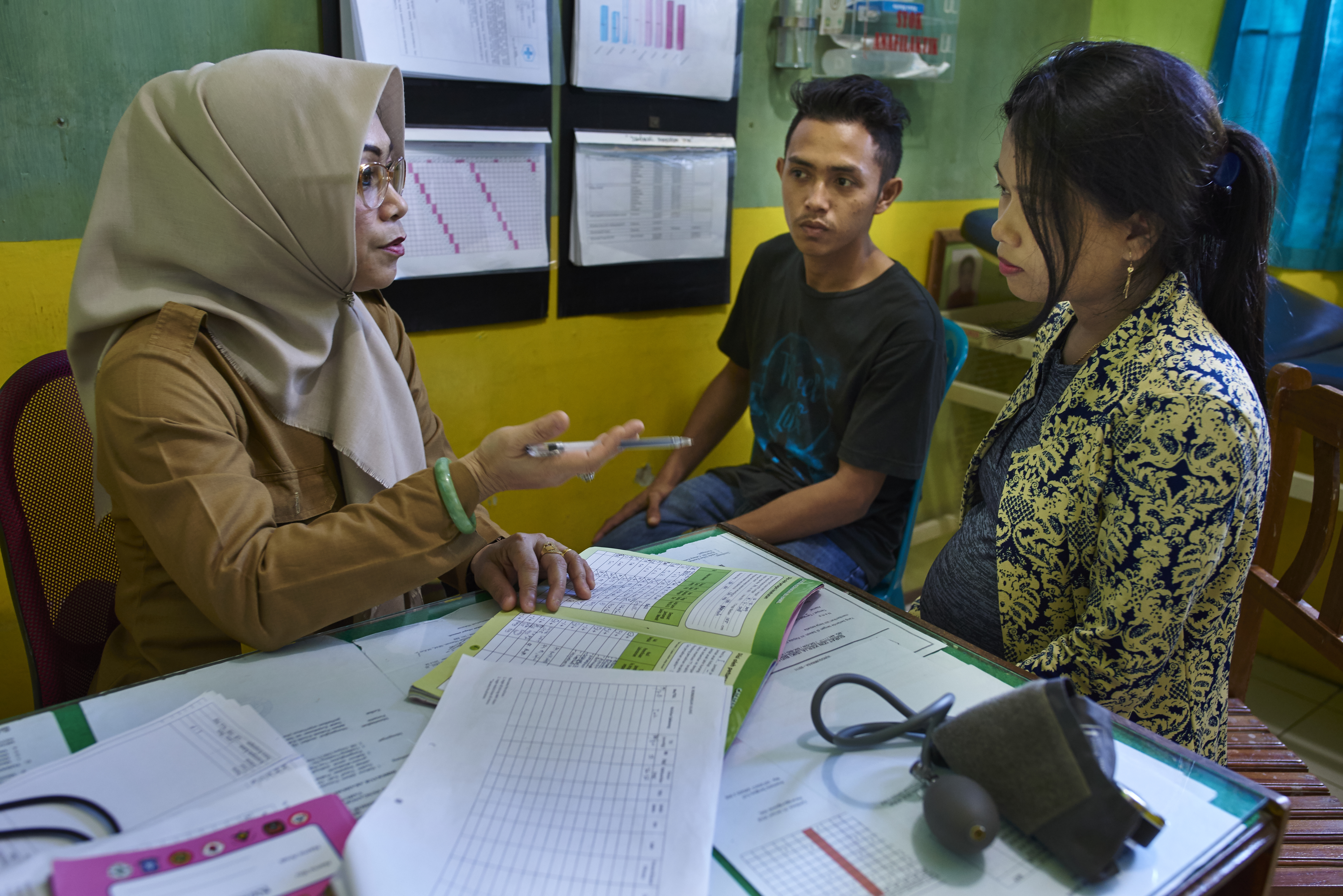 A female doctor talks to her female patient about her health condition. The patients partner sits in the background listening in on the conversation