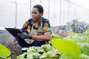 Woman smiling and writing on a clipboard in a greenhouse.