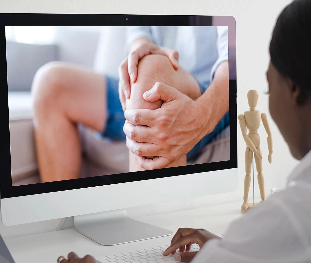 Um fisioterapeuta sentado em frente a um computador, visualizando a imagem de um paciente segurando o joelho com dor.