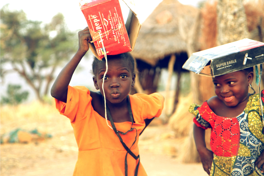 Two cheeky girls with cardboard boxes on their head. One in yellow dress is staring daringly at camera. Friend in red, blue and green patchwork dress is looking at her friend, giggling.