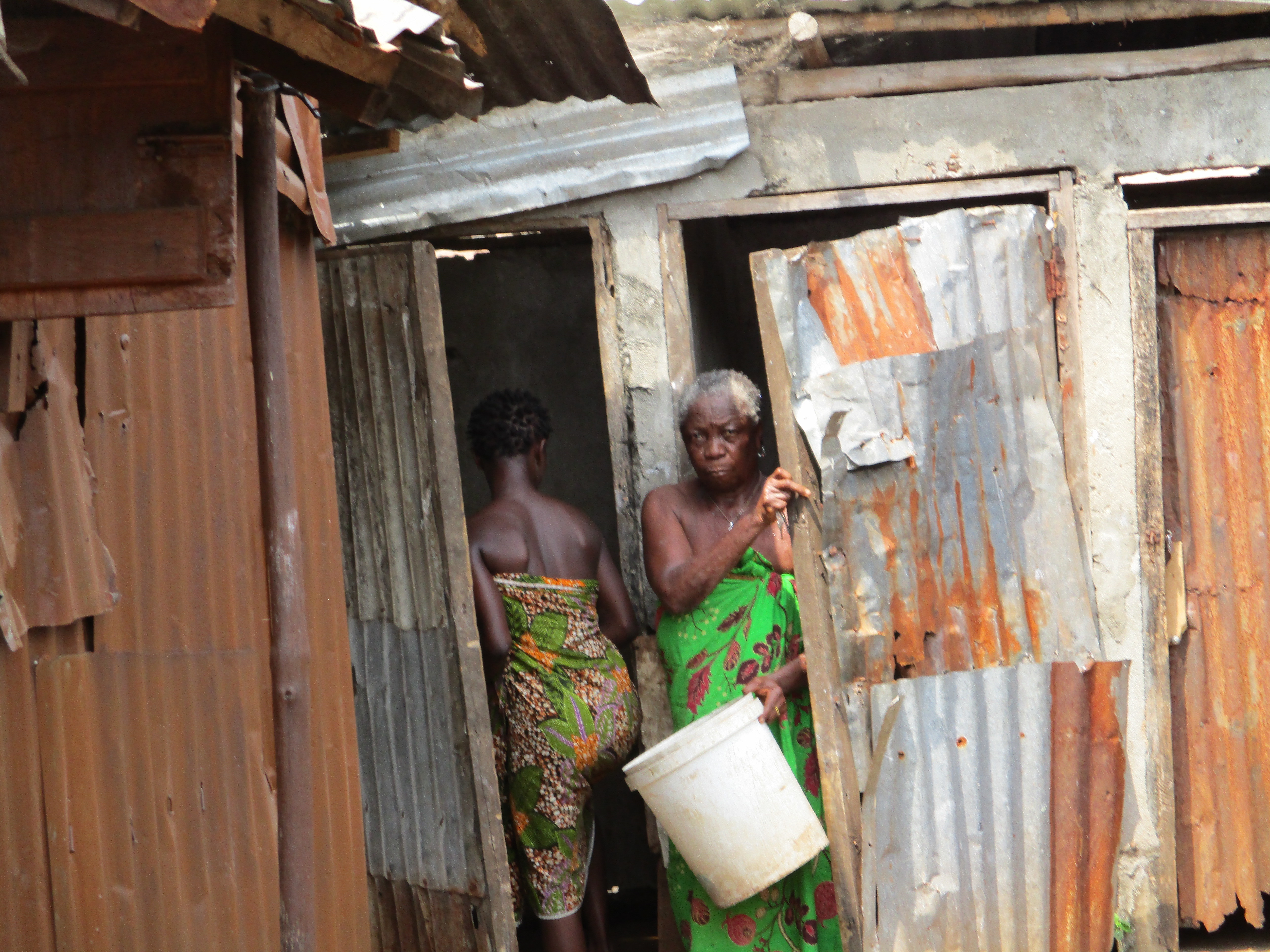 image shows a corrugated iron structure with two doors. one has a women walking in, and one has a woman leaving, holding a white bucket.