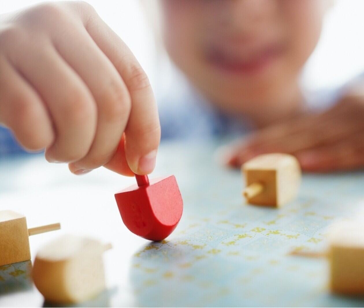 A close-up view of a child’s hand spinning a red wooden top on a tabletop, with several small wooden tops scattered nearby and the child’s face softly blurred in the background.