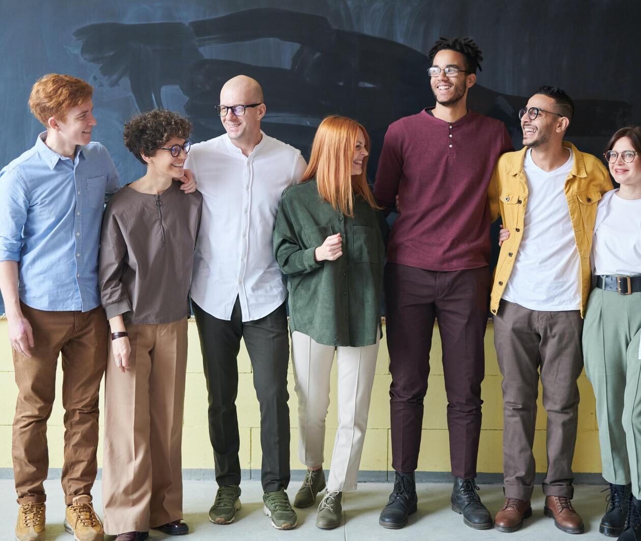 Seven people in business casual dress, a variety of heights, genders, ages and ethnicities stand in a line in front of a chalkboard. They are smiling, and have their arms around each other's shoulders and backs.