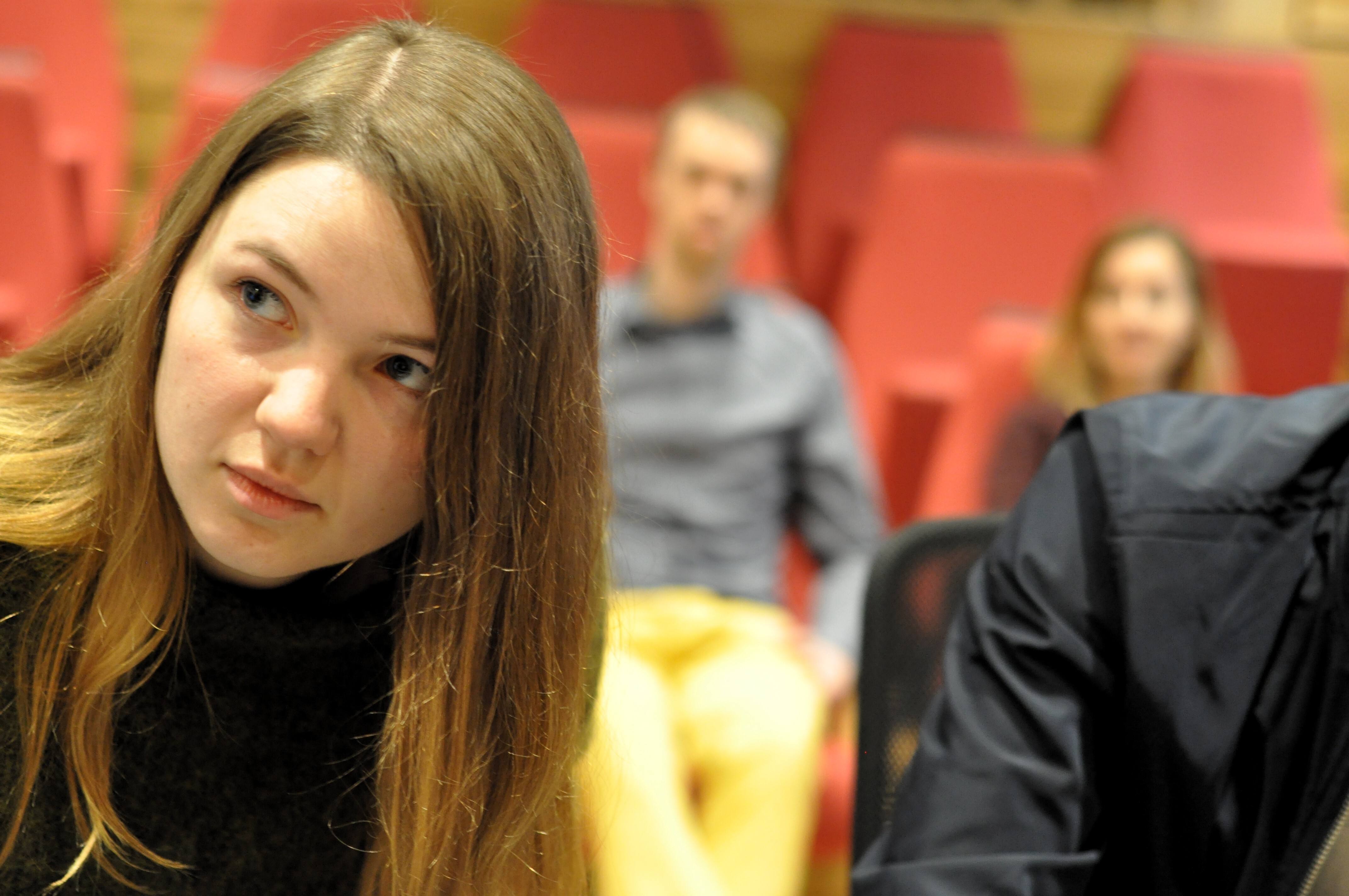 A woman is sitting in an auditorium, two people are sitting behind her. The background is blurred.