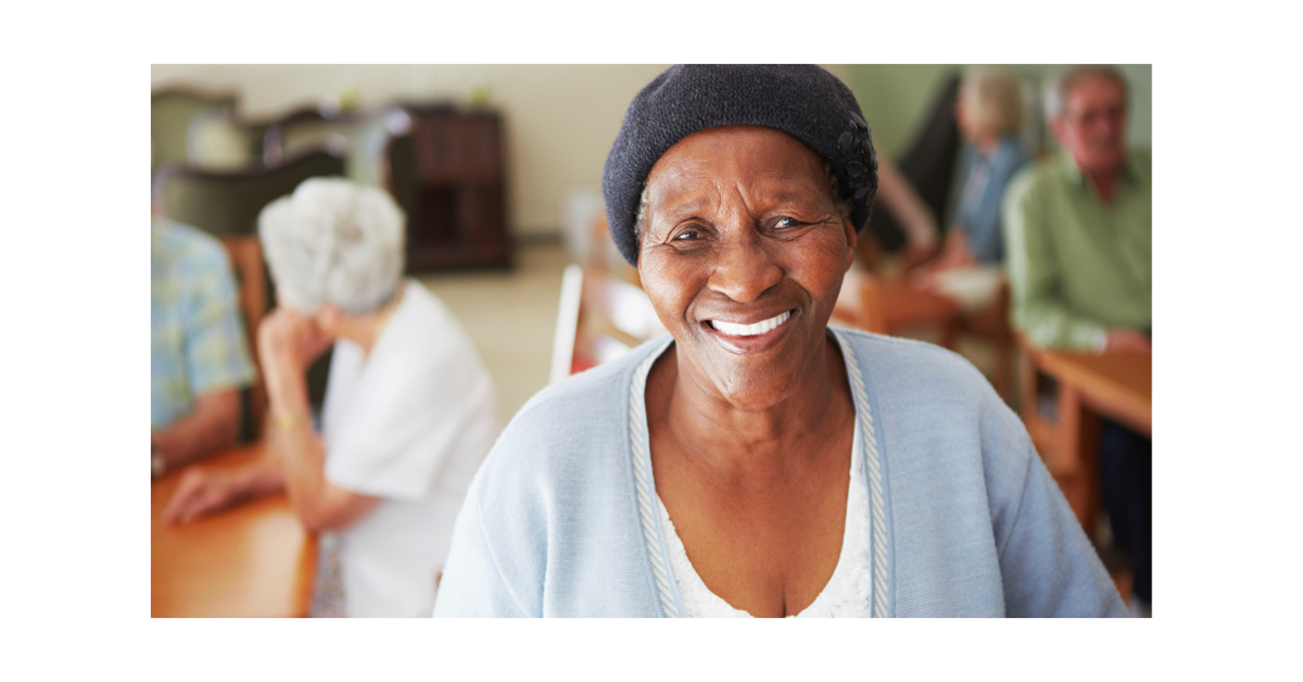 A picture of a lady smiling with other people in the background in what looks like a care home day room.