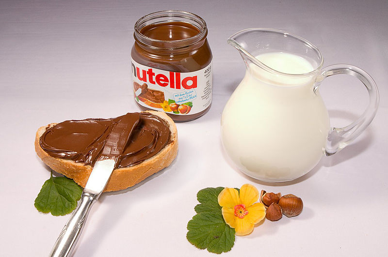 Nutella jar, nutella on bread with knife, glass of milk and hazelnuts.