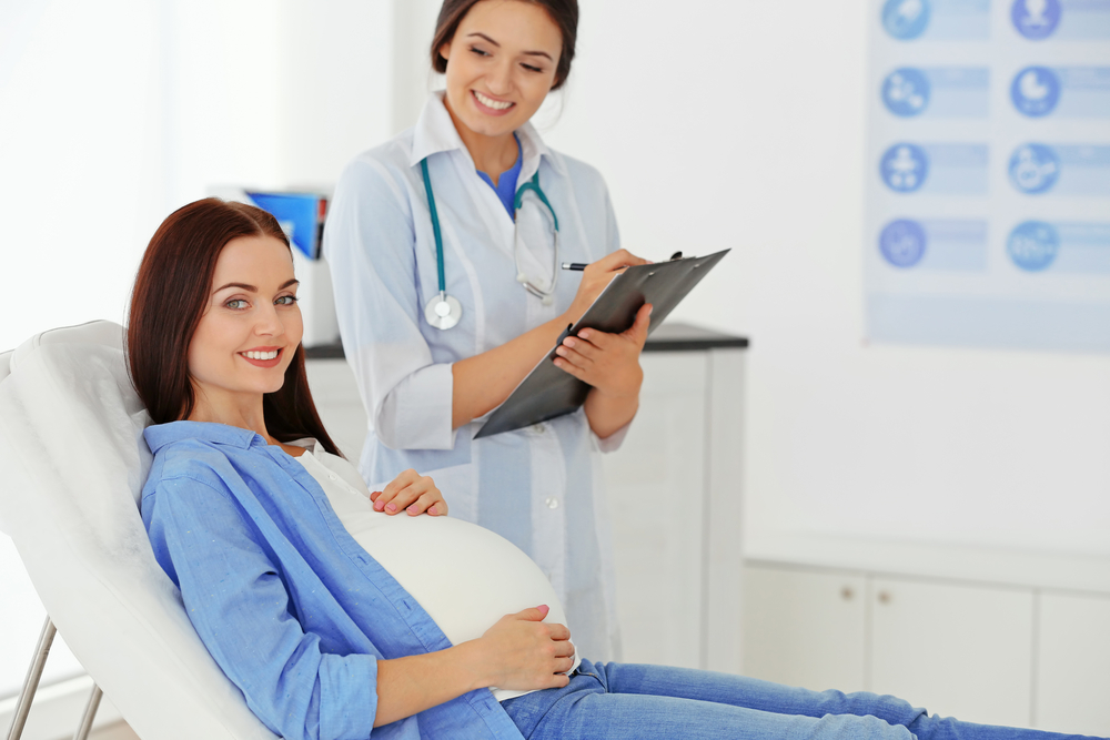 A young, pregnant mother reclines on an examination couch, while the health practitioner stands beside her with a clipboard, ready to take the details of the maternal history.