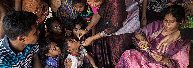 A woman gives oral medicine to a child surrounded by children and adult onlookers