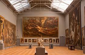 A gallery room from The Royal Museum of Fine Art, Brussels. Several people are in the room, which is large with high ceilings and wooden floors. The walls are covered in various art works, Renaissance in style but indistinguishable from the distance.