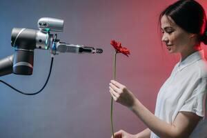 A robotic arm reaching out for a red flower held by a woman.