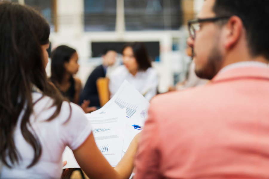 Man with pink jacket and woman with with t-shirt and long brown hair have backs to camera. They are looking together at financial documents with charts, and in the background are others seated and in discussion.