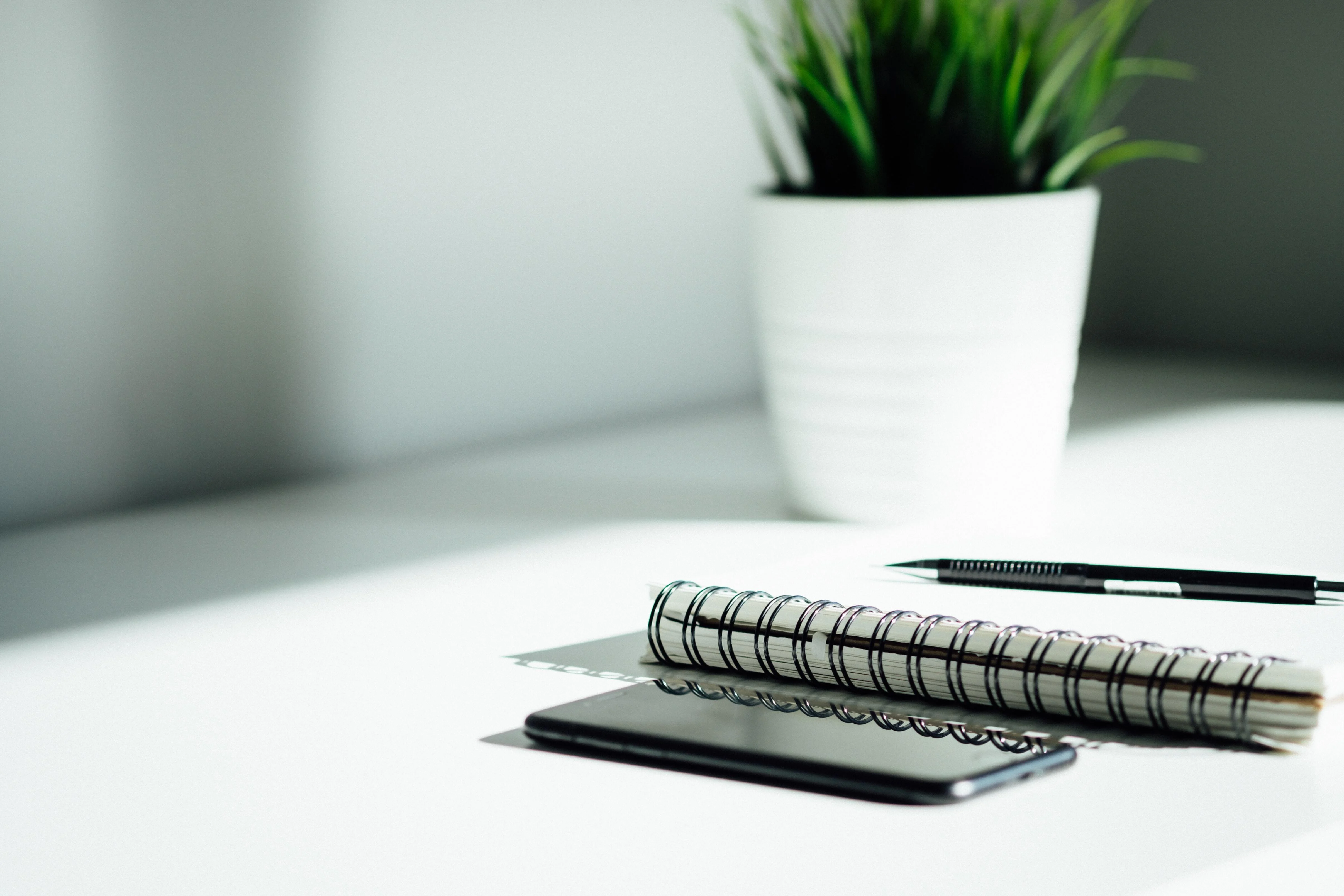 A notebook and pen, a mobile phone and a plant pot set on a white table