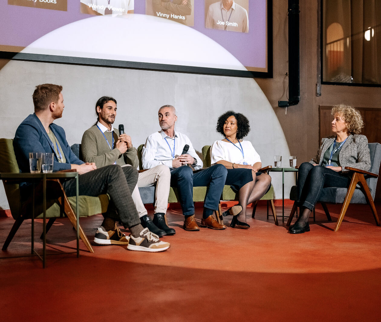 A panel of five people sitting in a row on a stage at a conference with a large presentation above their heads.